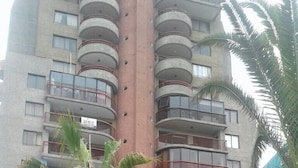 Un hotel de varios pisos con balcones, un pilar central rojo y un pequeño jardín con palmeras.
