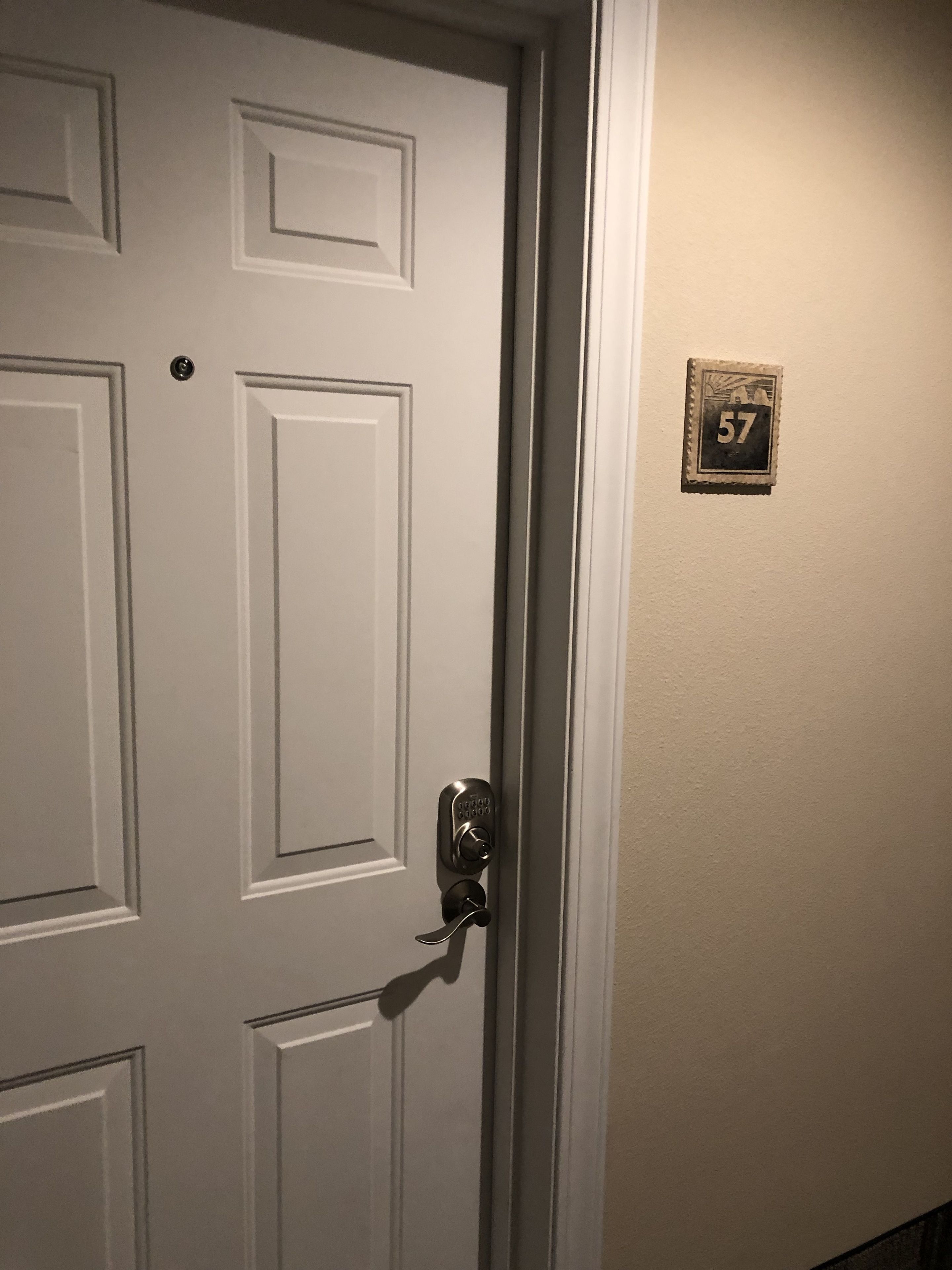 A white door with a keyhole and a doorbell. A small sign with the number 57 is visible on the wall to the right.