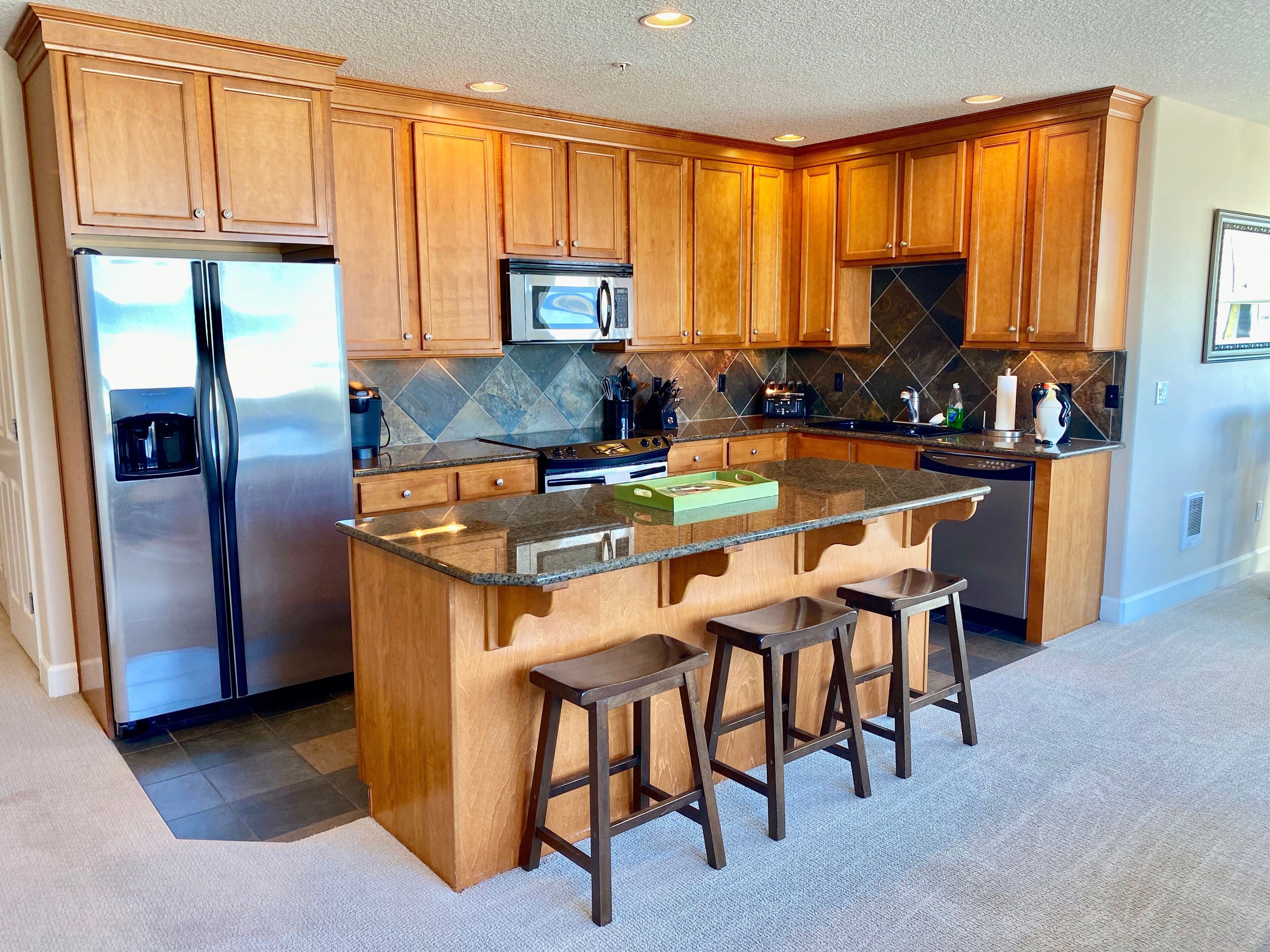 A kitchen with wooden cabinets, a stainless steel refrigerator, a microwave, and a granite countertop.