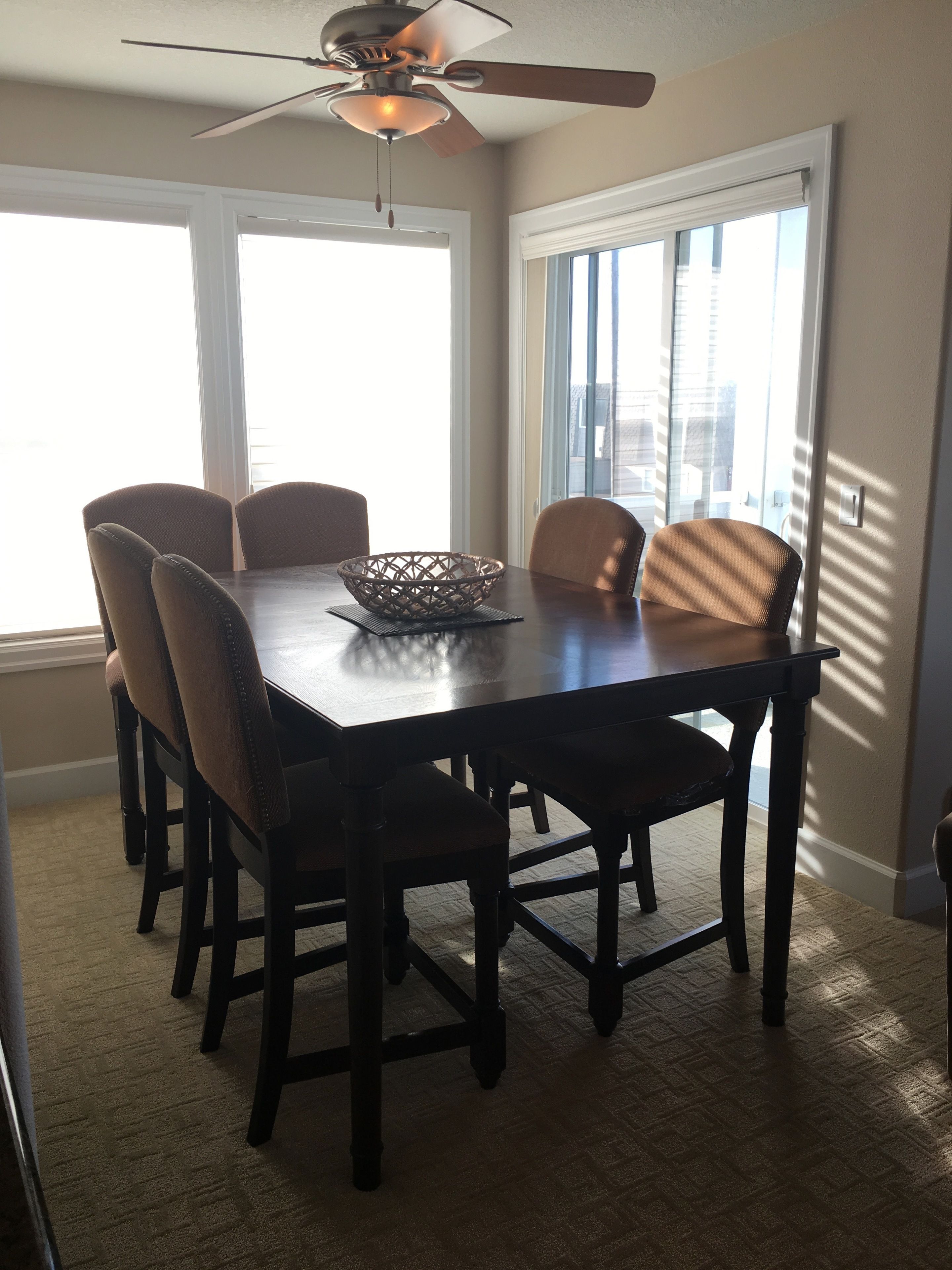 Comedor con una mesa de madera oscura, seis sillas, un ventilador de techo y ventanas grandes con persianas.