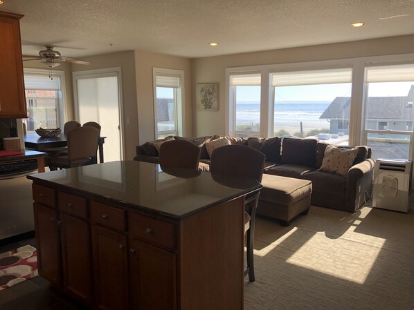 A spacious living area with a large window overlooking the beach, a cozy seating arrangement, and a kitchen area.