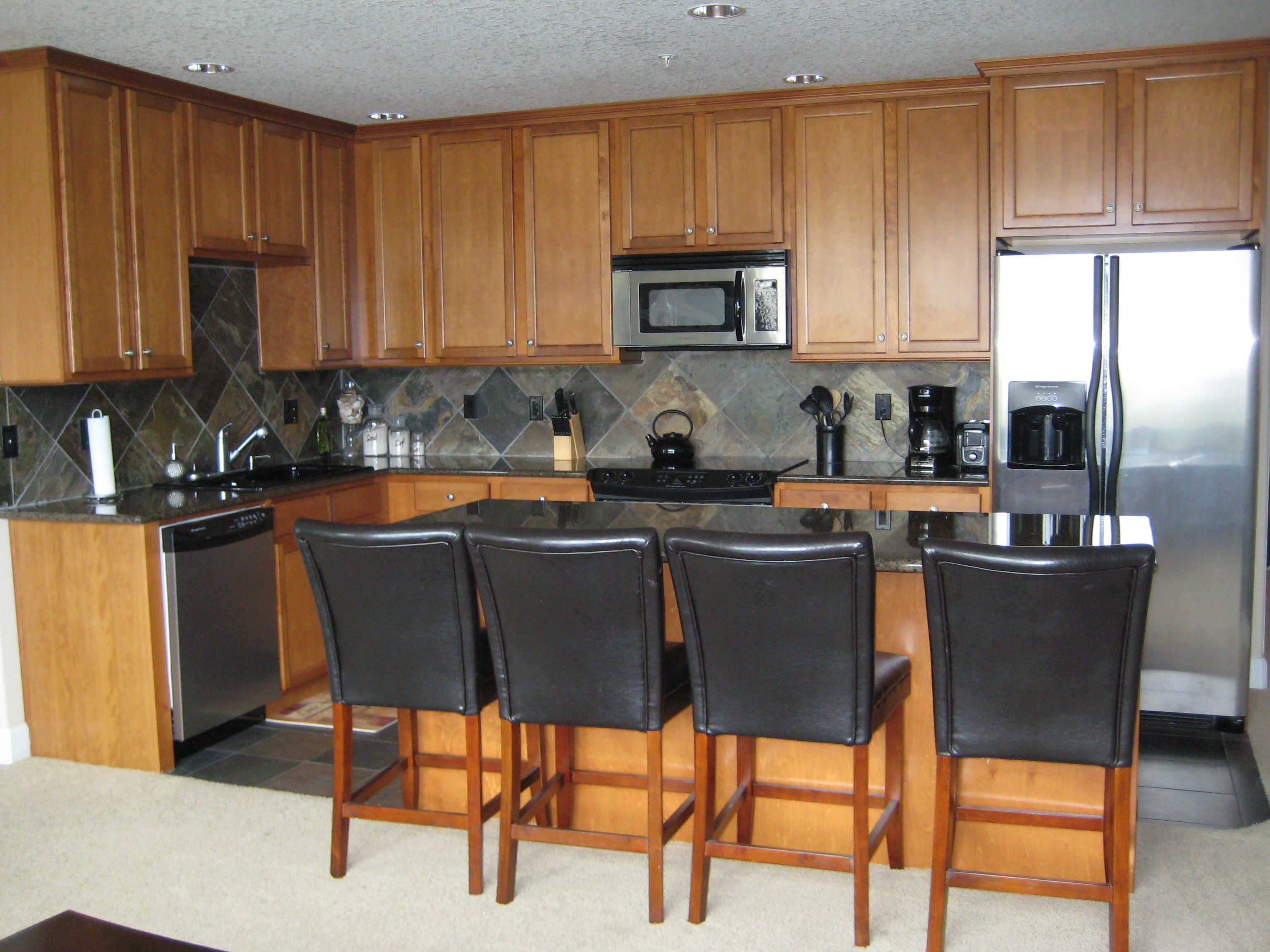 A kitchen with wooden cabinets, a stainless steel refrigerator, a microwave, a coffee maker, and four black chairs.