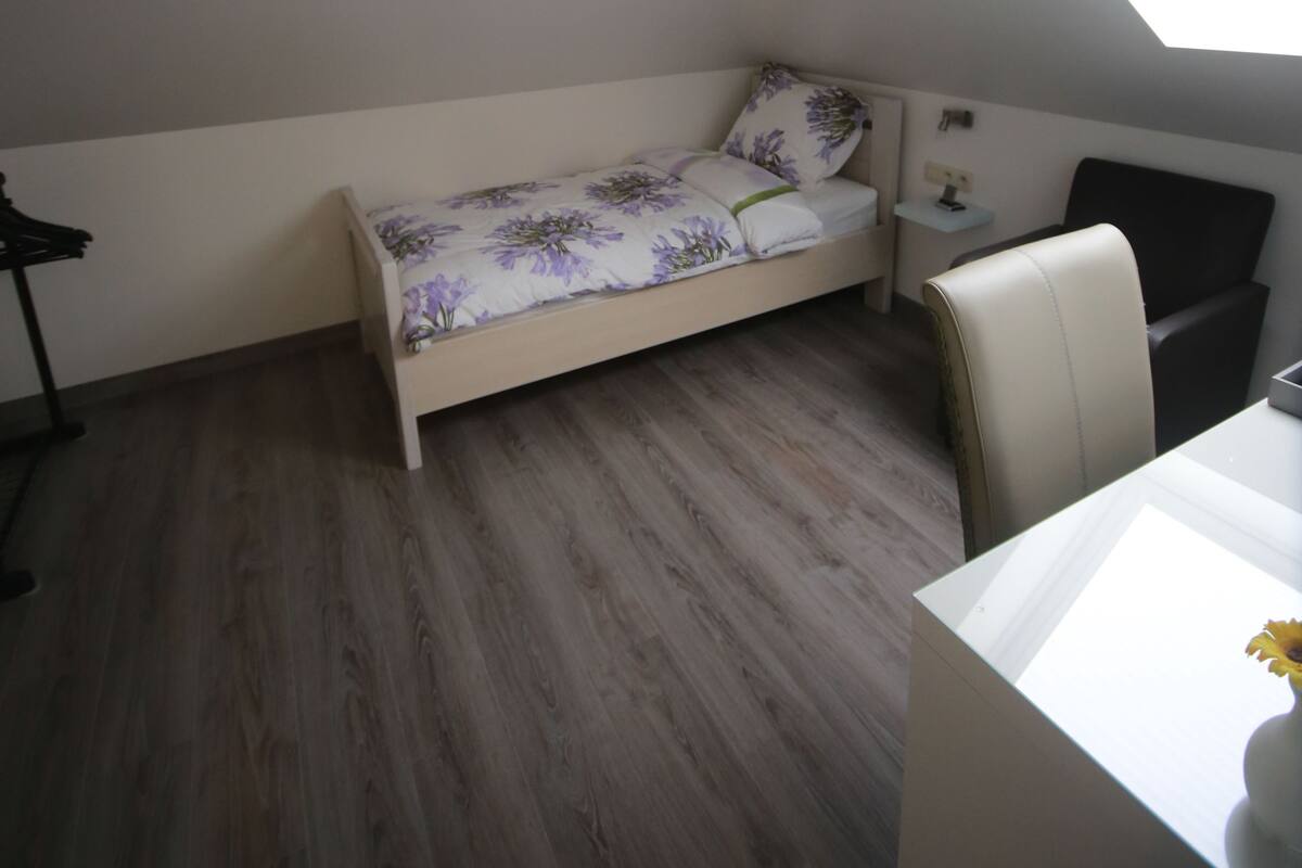 A small, single-bed room with a sloped ceiling and skylight, wooden flooring, a bedside table, and a chair.