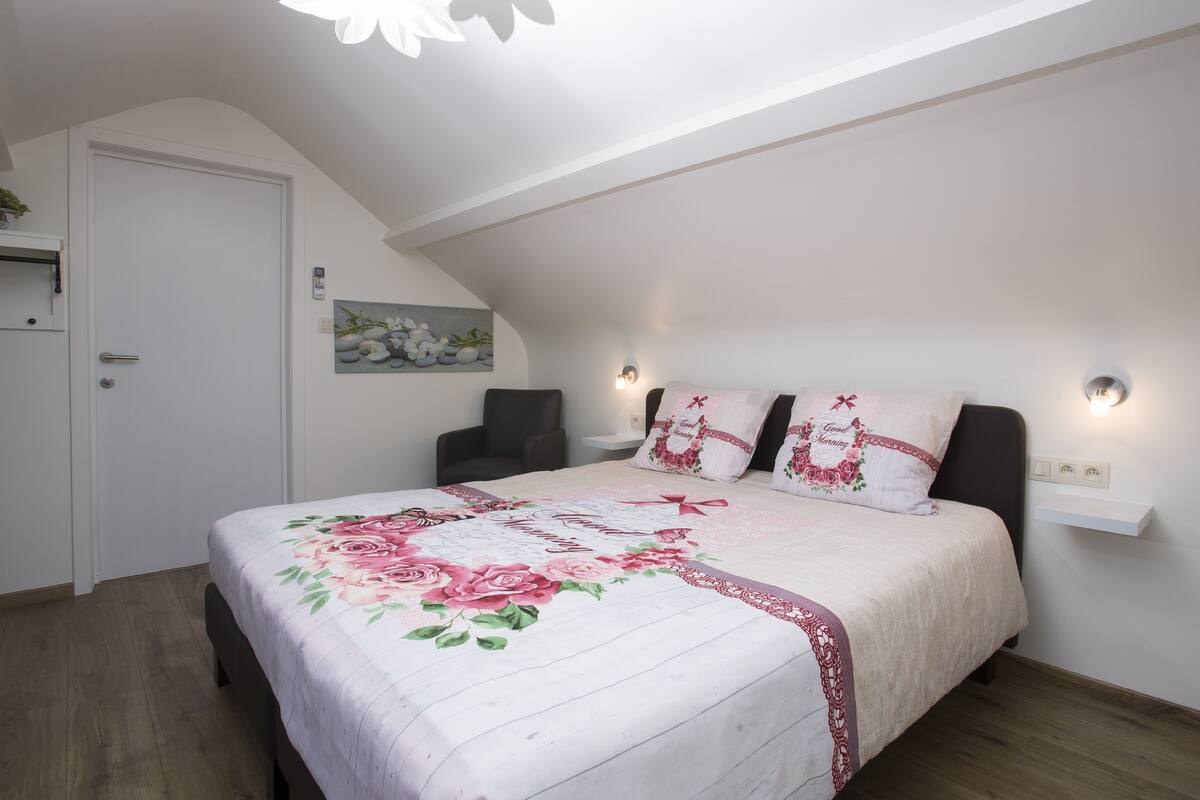 A bedroom with a bed, a nightstand, a wall-mounted shelf, a door, and a ceiling light fixture.