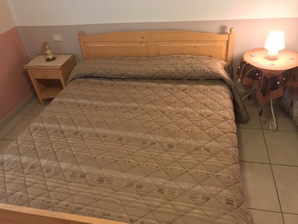 Ein Bett mit gesteppter Bettdecke, ein hölzernes Kopfende, ein Nachttisch mit Lampe und ein kleiner Tisch ebenfalls mit Lampe.