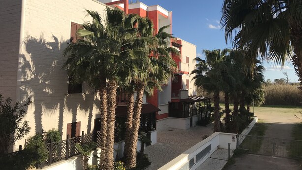 A building with a red and white facade, palm trees, and a paved courtyard. - Residence Massilor (Gallipoli)