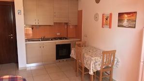 A compact kitchen with a gas stove, sink, and oven. There is a dining table with chairs and a small rug. The room has a tiled floor and a door leading to another area. - Residence Massilor (Gallipoli)