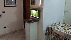 A room with a television on a stand, a clock on the wall, a framed picture, and a coffee table with a coffee-themed cover. - Residence Massilor (Gallipoli)