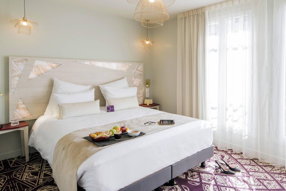 A hotel room with a large bed, two bedside tables, a bedside lamp, a fruit basket, and a pair of shoes on the floor.