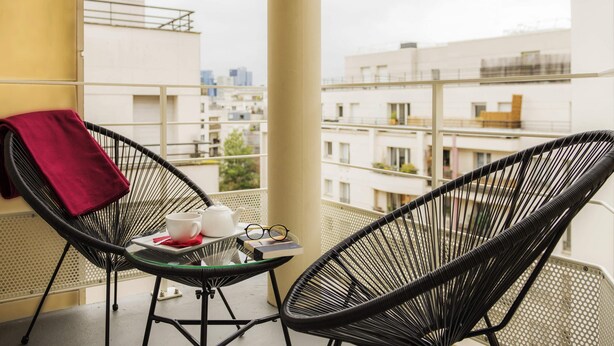A balcony with two black wicker chairs, a small table with a teapot, cups, and a book, and a red towel hanging on the chair. - Hôtel Mercure Paris Pont de Levallois Neuilly (Levallois-Perret)