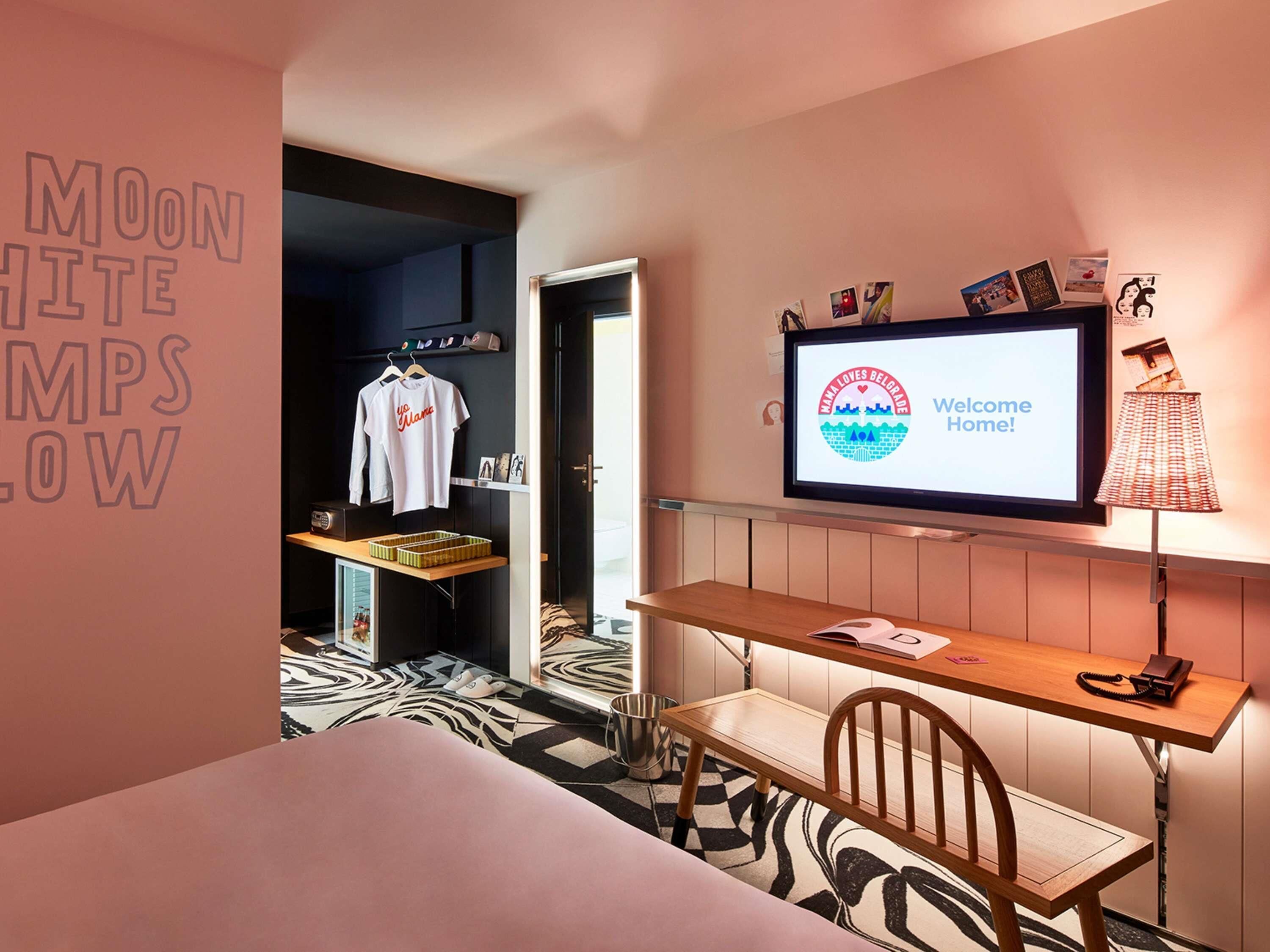 A hotel room with a bed, a desk with a chair, a TV displaying 'Welcome Home!', and a wall with a sign reading 'MOON WHITE MPS LOW'.
