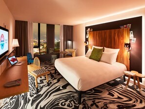 A hotel room with a bed, a desk with a laptop, a chair, a small table, and a view of the cityscape. - Mama Shelter Belgrade (Belgrade)