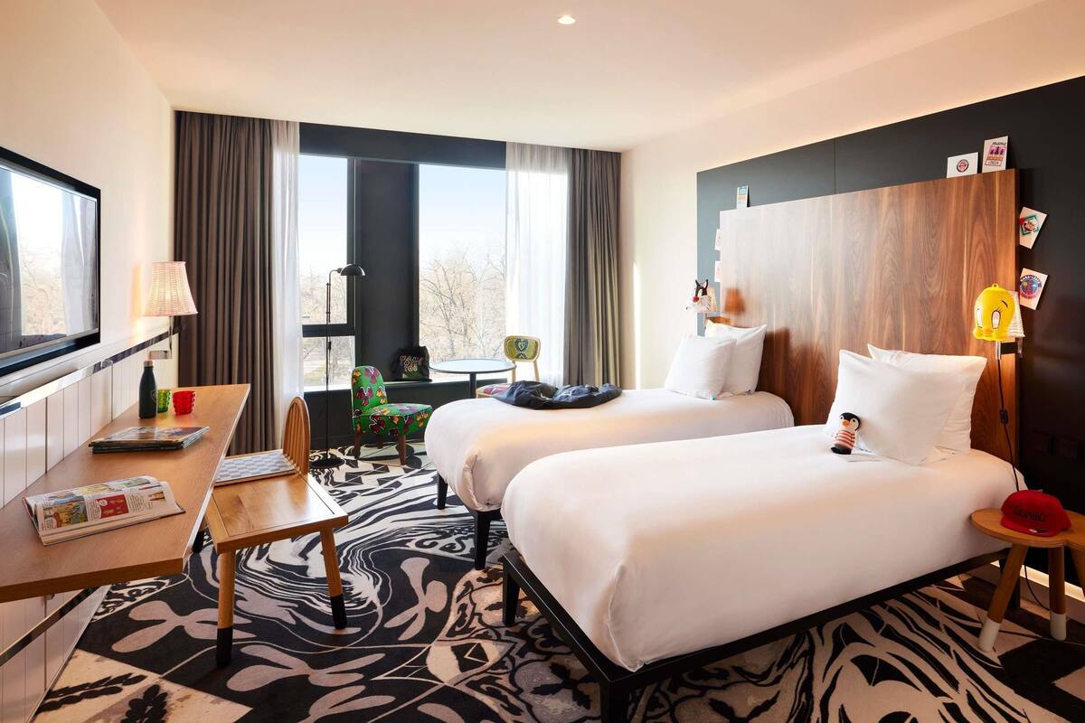 Ein Hotelzimmer mit zwei Betten, einem Schreibtisch, einem Fernseher, einem Fenster mit Vorhängen und Blick auf Bäume.