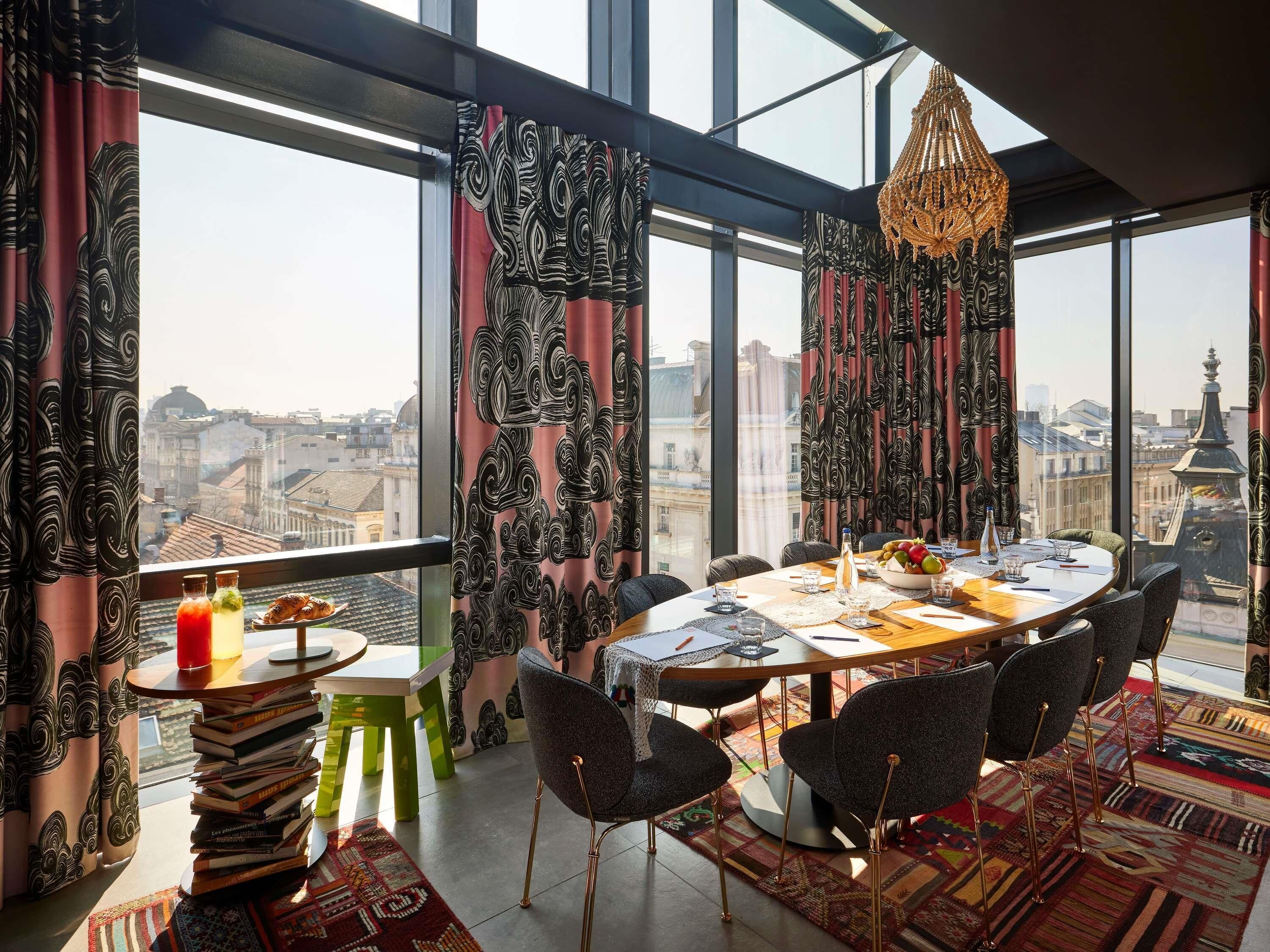 Un comedor con una mesa preparada para cuatro personas, una araña y vistas de la ciudad a través de amplias ventanas.