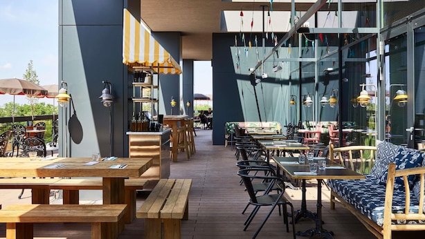 An outdoor dining area with wooden tables and chairs, a bar area with a striped awning, and hanging colorful flags. - Mama Shelter Belgrade (Belgrade)