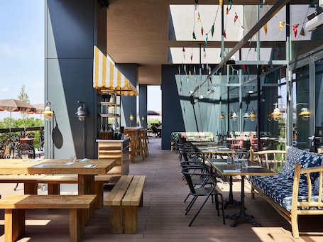 An outdoor dining area with wooden tables and chairs, a bar area with a striped awning, and hanging colorful flags.. Mama Shelter Belgrade