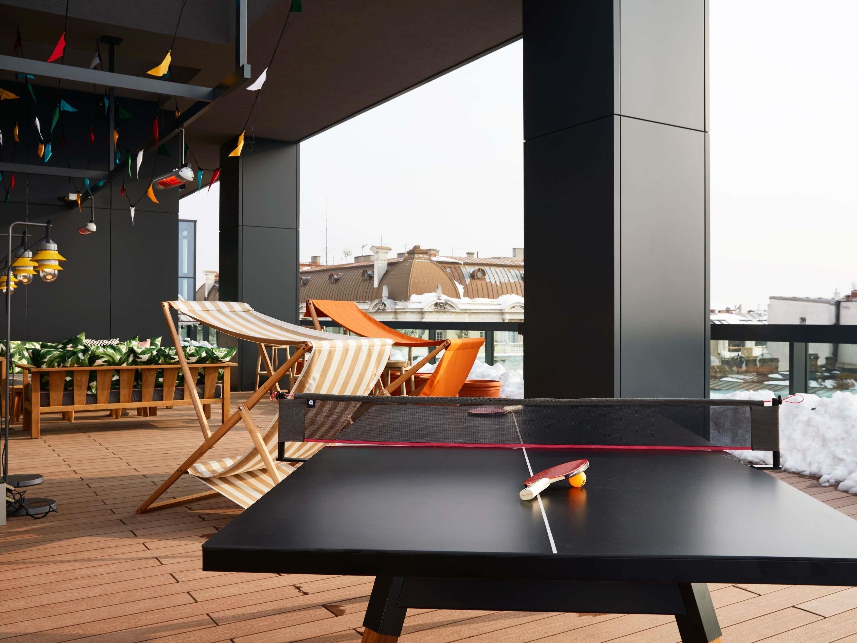 Una mesa de ping pong con una raqueta y pelota, dos sillas de playa a rayas y un banco de madera con plantas.