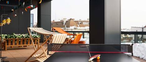 Una mesa de ping pong con una raqueta y pelota, dos sillas de playa a rayas y un banco de madera con plantas.