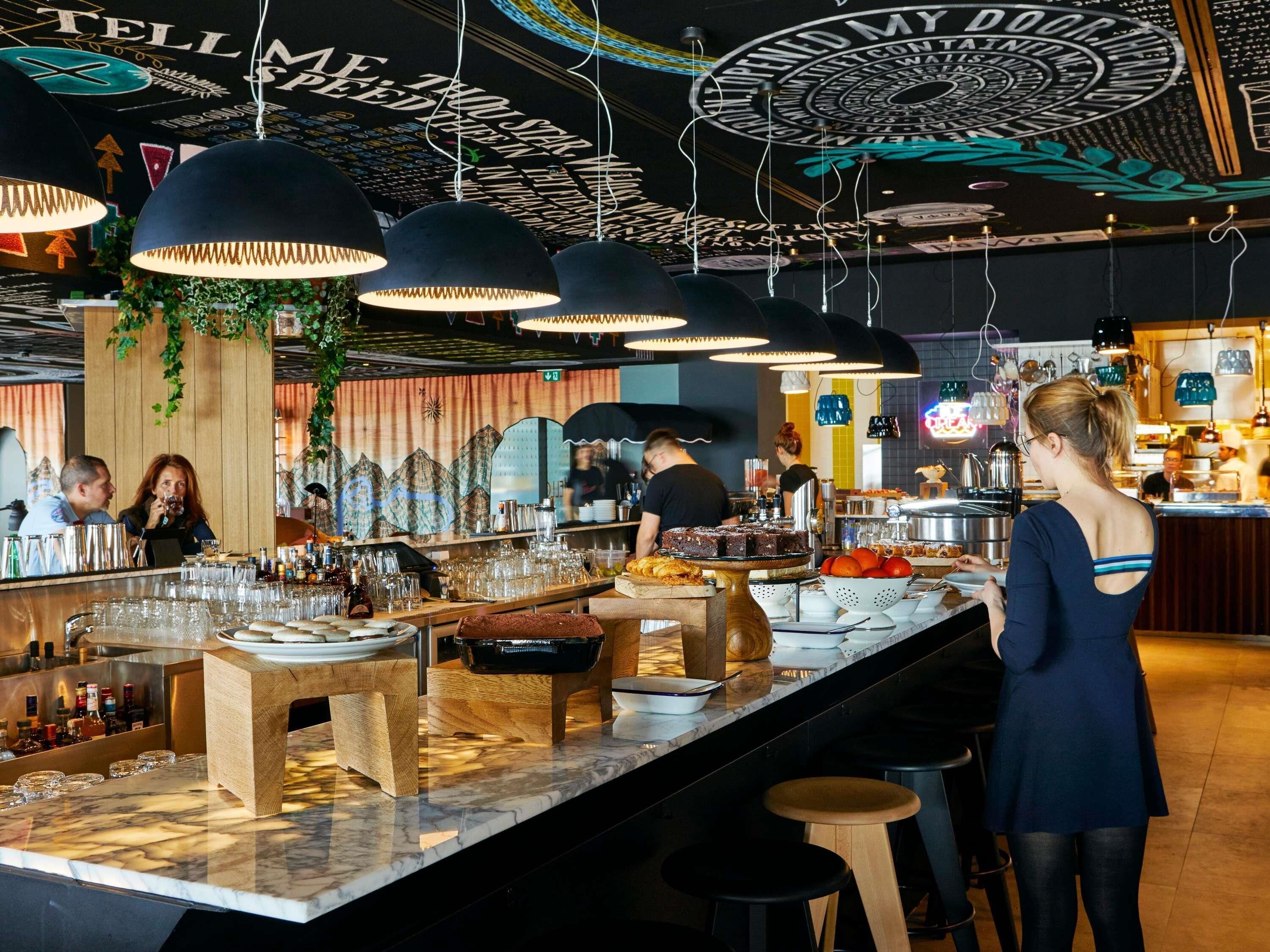 A bar area with multiple hanging pendant lights, a counter with glasses and bottles, and a person serving.