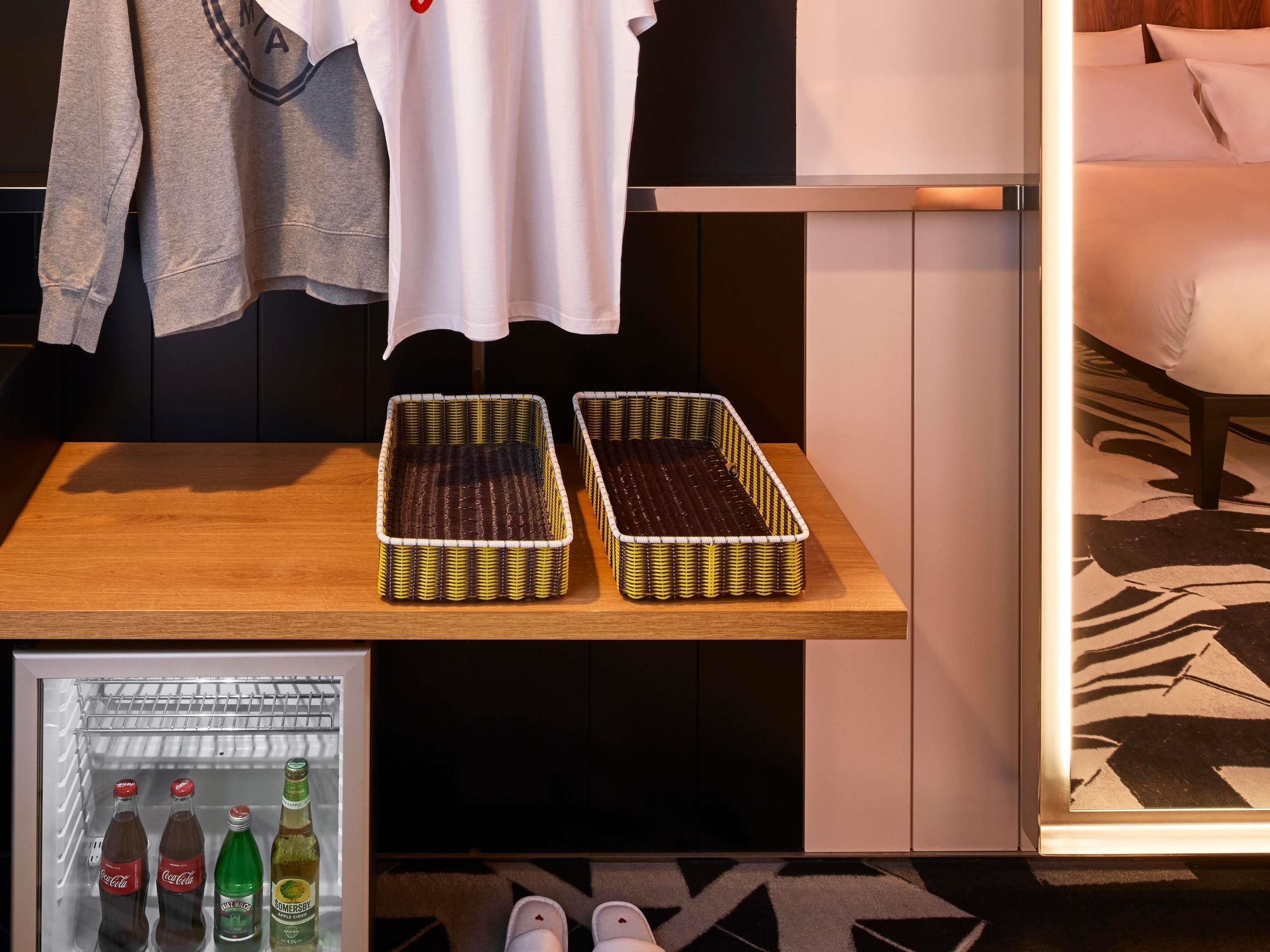 A room with a wooden table, two wicker baskets, a refrigerator with beverages, and a bed with a patterned rug.