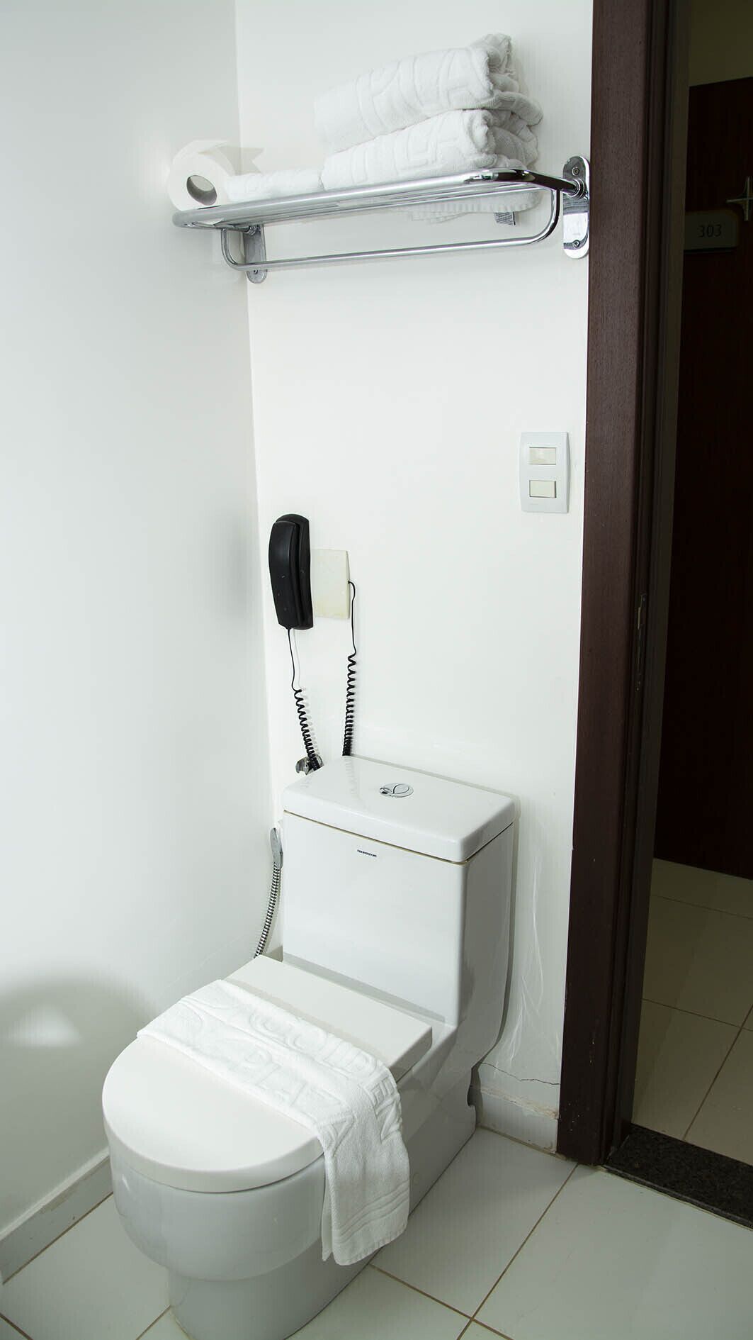 A white toilet with a towel on the seat, a telephone mounted on the wall, and a towel rack above the toilet.