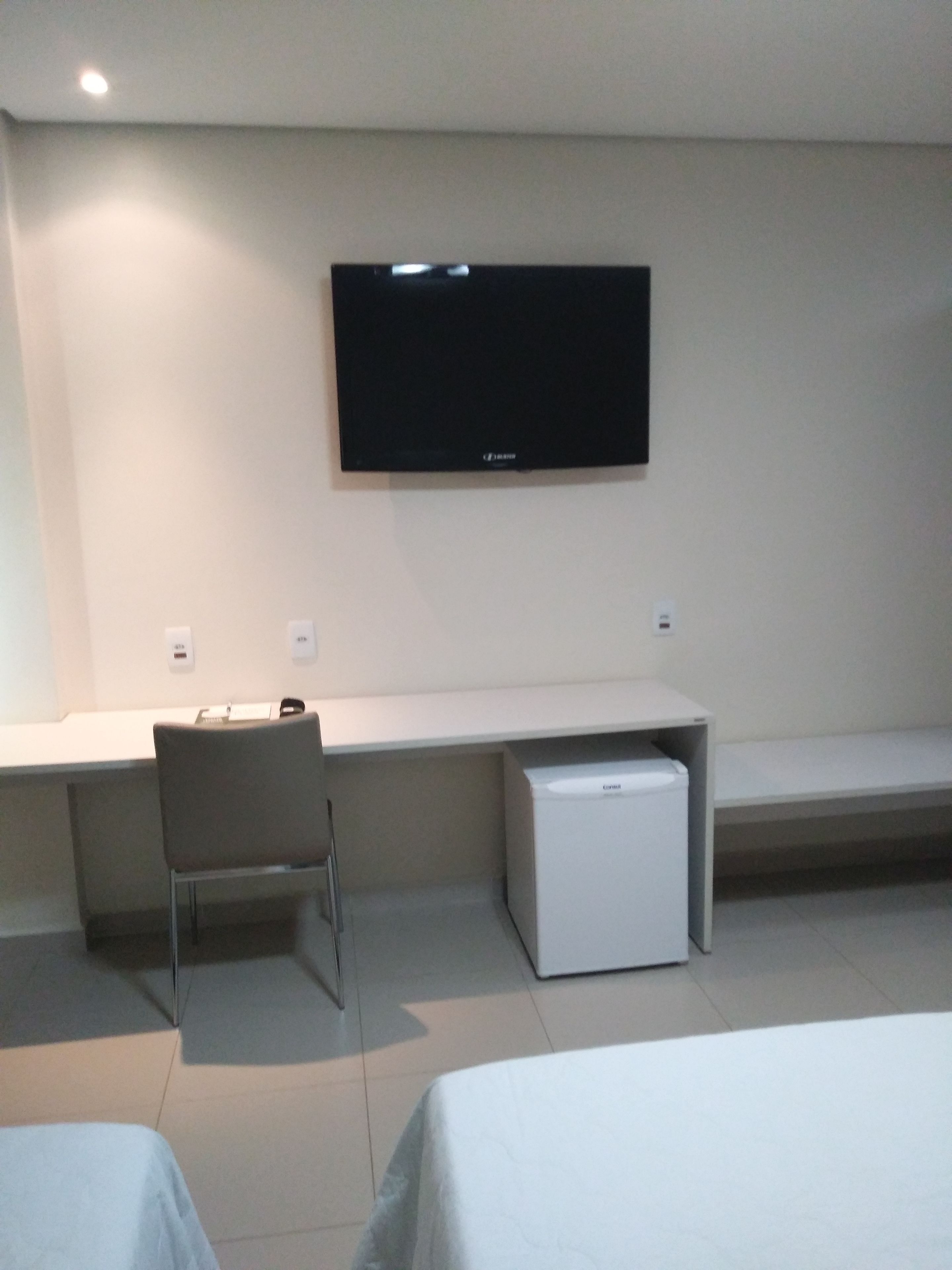 A hotel room with a bed, a desk with a chair, a television mounted on the wall, and a small refrigerator.