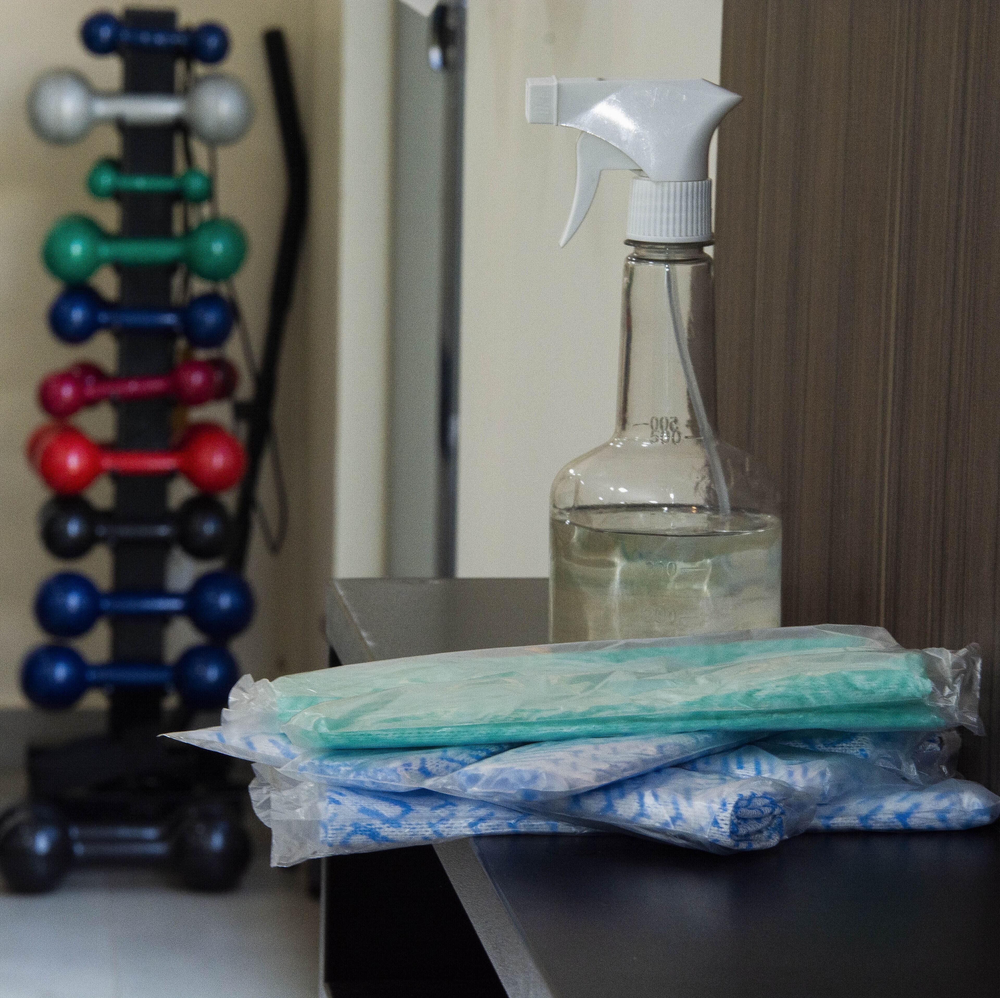 A table with a spray bottle, a pack of tissues, and a dumbbell rack in the background.