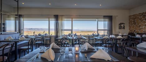 A restaurant with a long table set for dining, large windows offering a view of the desert, and a stone wall on one side.
