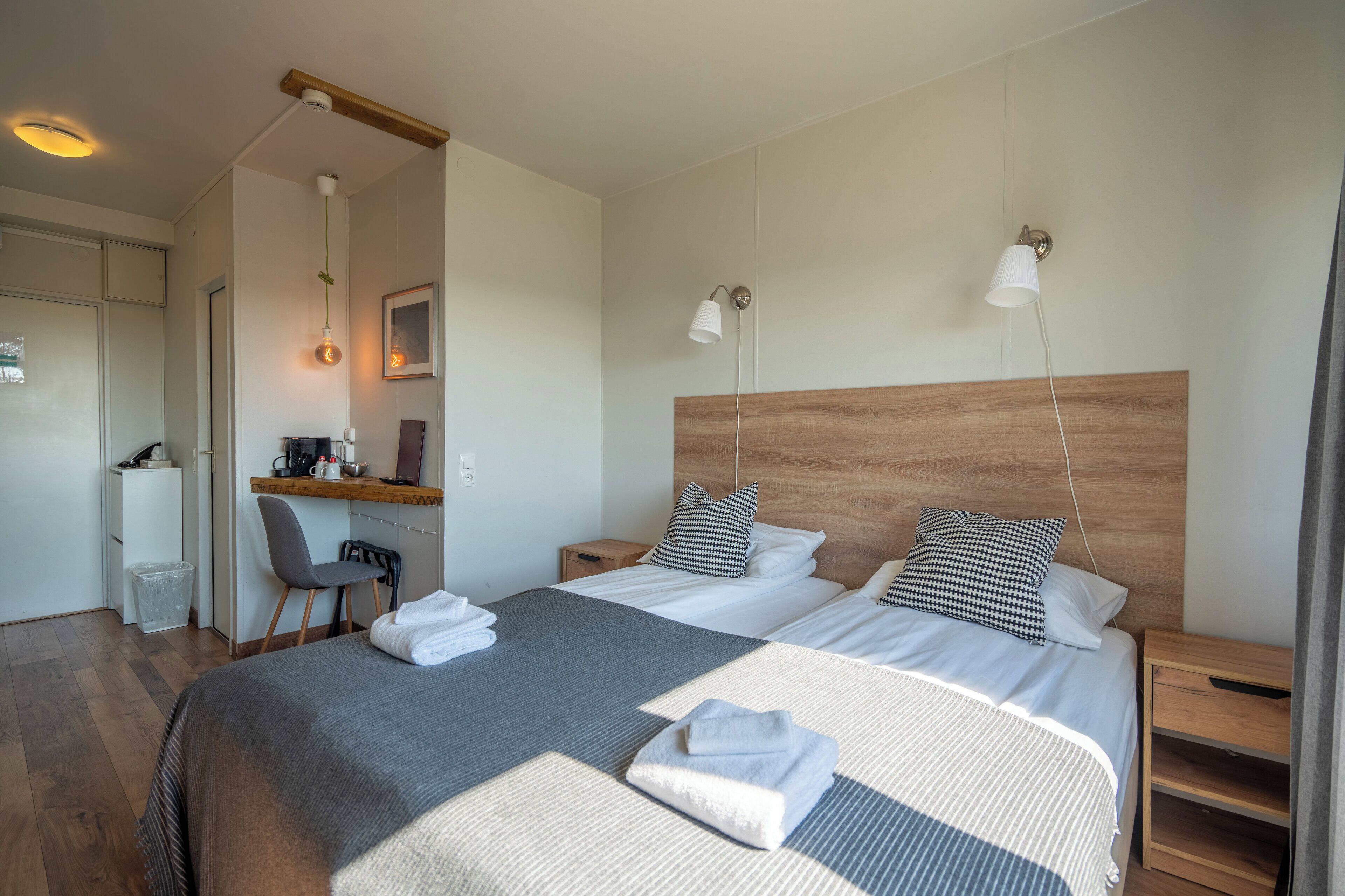 Une chambre d’hôtel avec un grand lit, deux tables de chevet, un bureau avec une chaise, une petite poubelle et une tête de lit en bois.