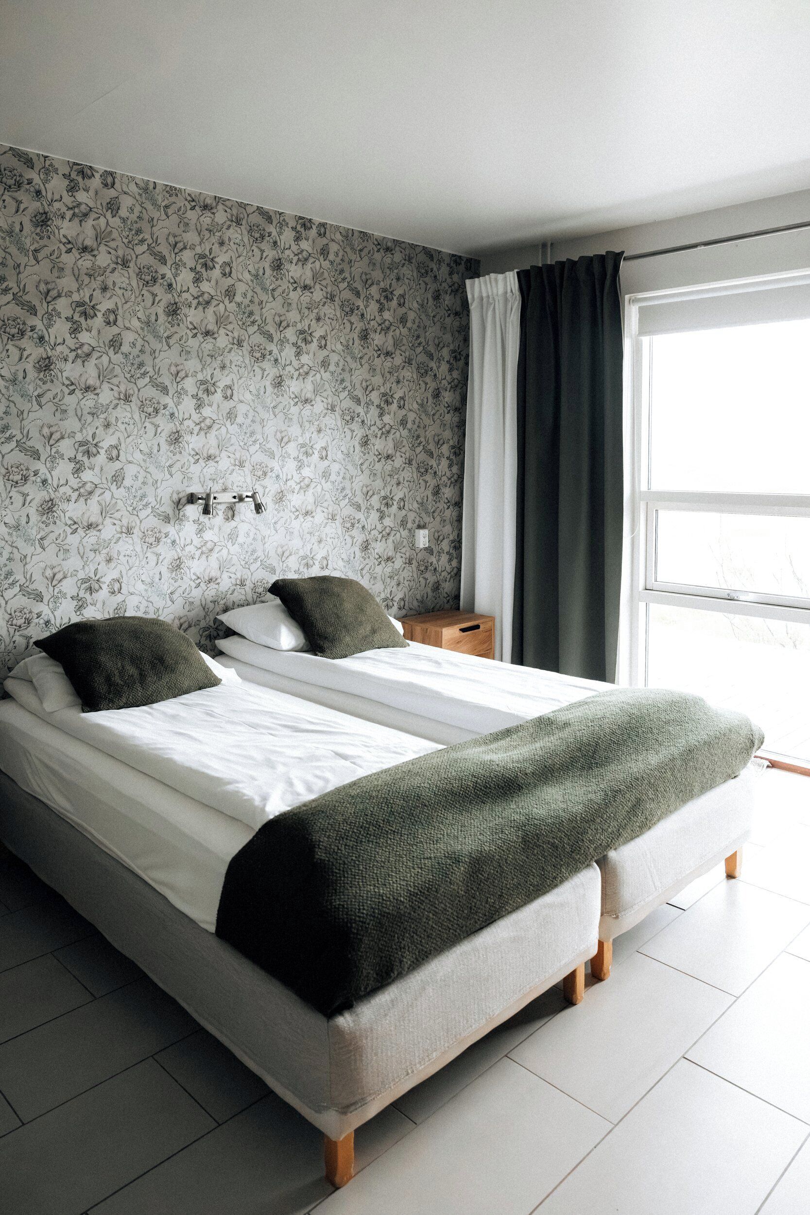 Een tweepersoonsbed met een groene deken, een nachtkastje met een lamp en een raam met gordijnen.