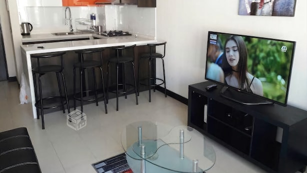 A modern kitchen with white and orange cabinets, a black countertop, and a glass dining table with four black bar stools. A flat-screen TV is mounted on the wall. - Depto en Iquique Ed. Terranova 1108 (Iquique)