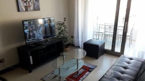 A living room with a TV, a glass coffee table, a black sofa, and a sliding glass door leading to a balcony. - Depto en Iquique Ed. Terranova 1108 (Iquique)