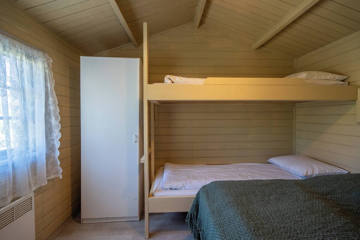 Ein Schlafraum mit Etagenbetten, Holzboden und -wänden, einer weißen Tür und einem Fenster mit Spitzenvorhängen.