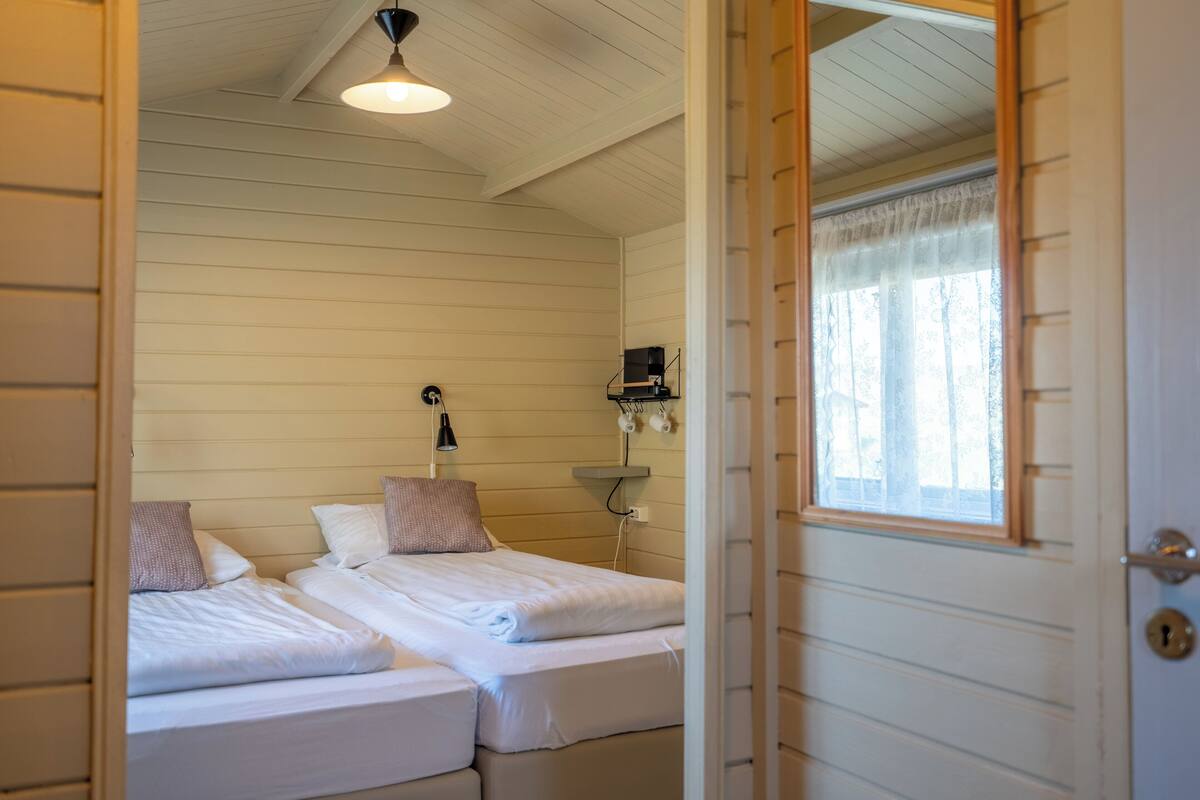 Ein Zimmer mit zwei Betten, einer Deckenleuchte, einem Wandregal mit Lampe und einem Fenster mit Vorhängen.