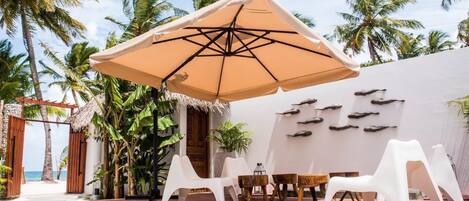 A beachside lounge area with a thatched-roof entrance, white lounge chairs, a table, and a large umbrella.