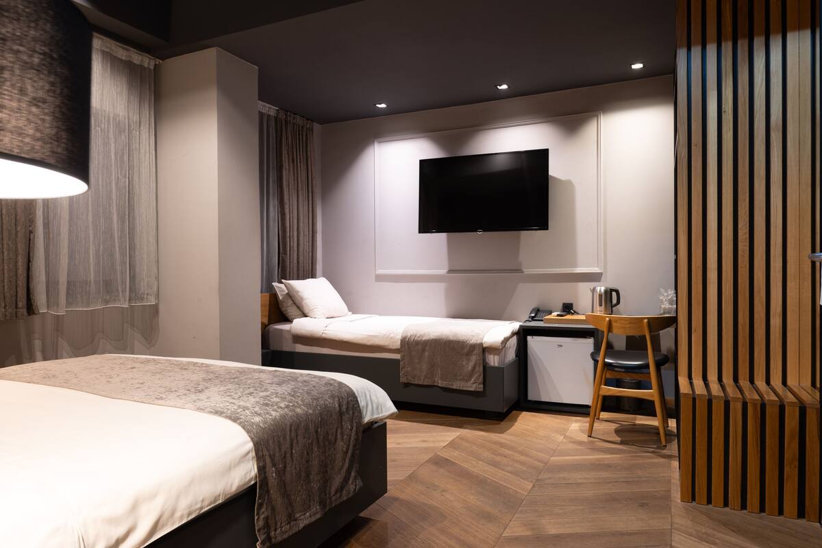 A modern hotel room with two beds, a flat-screen TV mounted on the wall, a small desk with a chair, and a wooden partition.