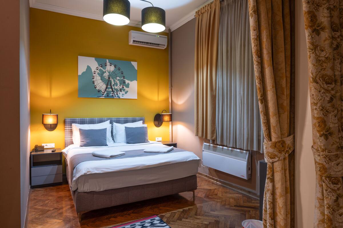 A hotel room with a bed, bedside lamps, a wall-mounted air conditioner, and a window with curtains.