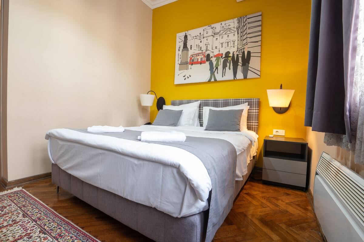 A neatly made bed with a grey and white duvet, a small bedside table with a lamp, and a wall-mounted light.