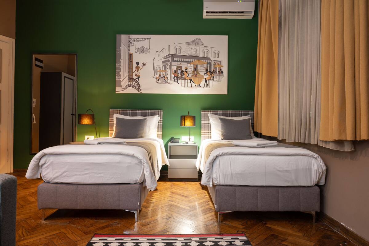 A hotel room with two beds, a green wall, a painting, and a window with curtains.