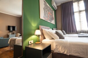 Habitación de hotel con una pared verde decorativa, una cama grande con ropa blanca, una mesita de noche con lámpara, un televisor de pantalla plana y una ventana con cortinas.