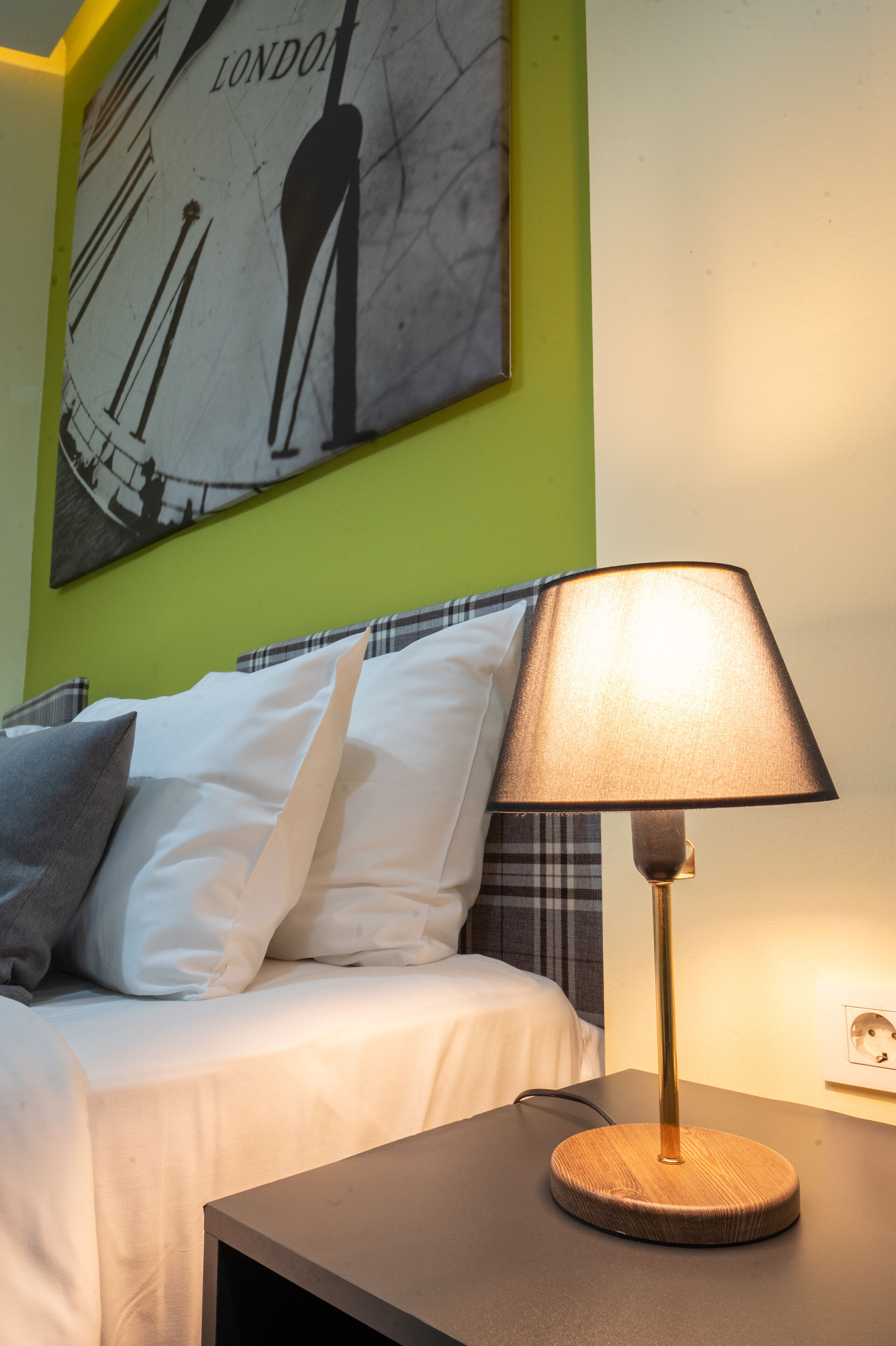 A neatly made bed with white bedding and a plaid headboard, a bedside table with a lamp, and a large wall clock with the word "LONDON" on it.