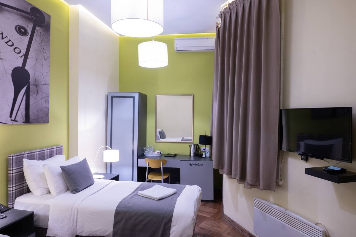 Quarto de hotel com cama, escrivaninha com cadeira, televisão, espelho e janela com cortinas.