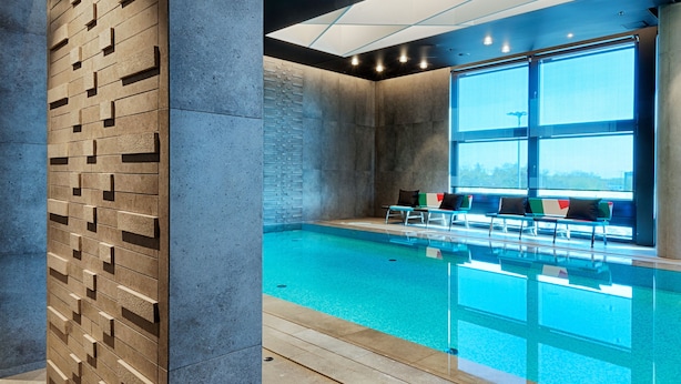 A modern indoor swimming pool with a clear view of the sky through large windows, a wooden wall with geometric patterns, and a seating area with chairs and cushions. - Renaissance Warsaw Airport Hotel, an Marriott International (Warsaw)