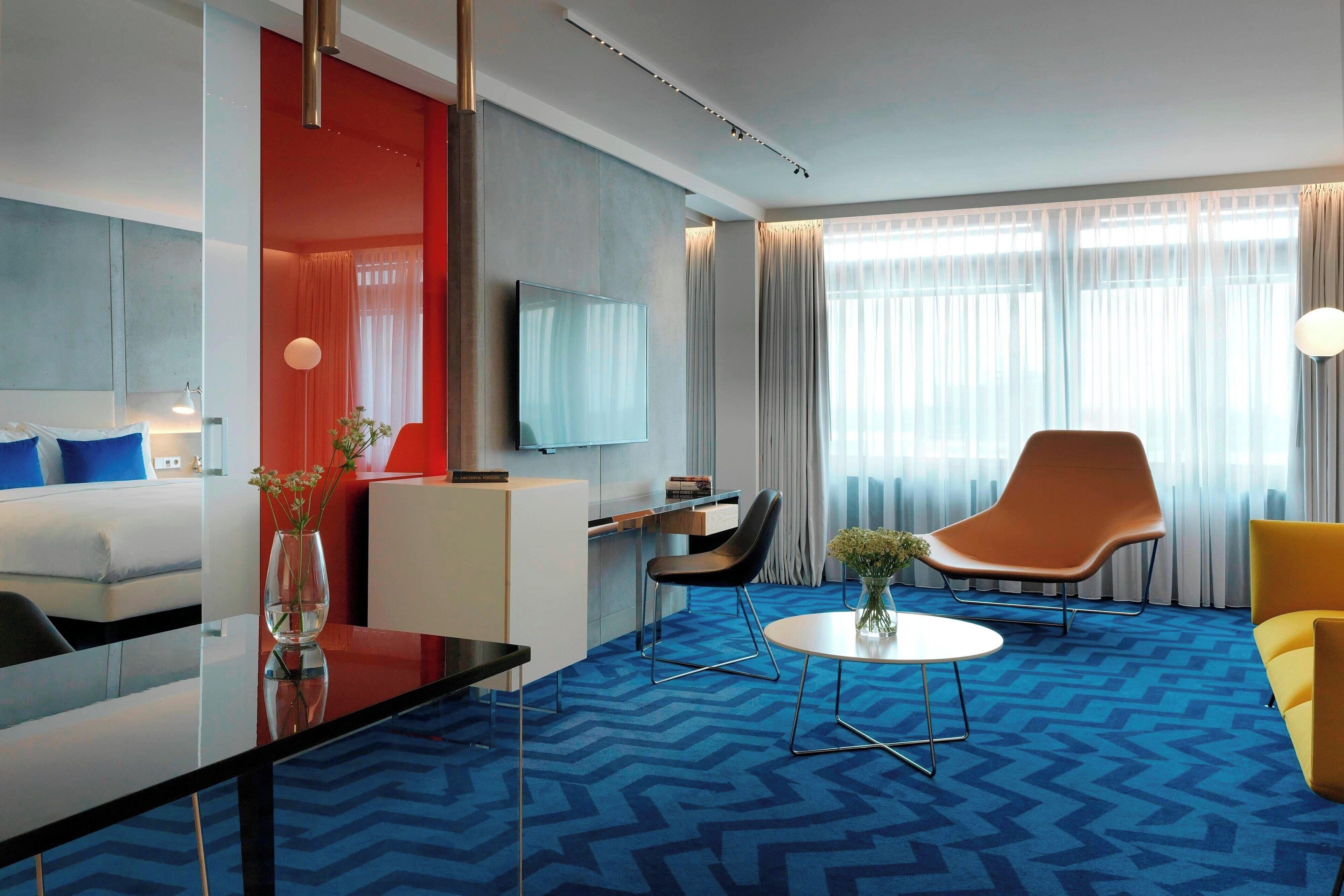 Une chambre d’hôtel moderne avec une moquette bleue, un téléviseur à écran plat fixé au mur, un canapé, un fauteuil et une petite table sur laquelle est posé un vase de fleurs.