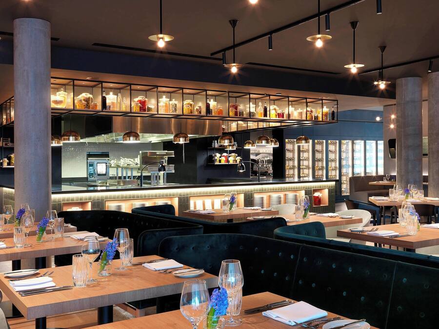 Restaurante moderno com mesas de madeira, assentos estofados em verde escuro e um bar com vitrines de vidro.