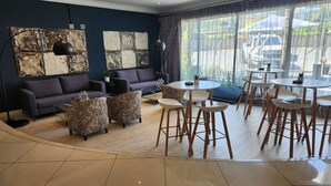 A modern hotel room with a dark blue accent wall, a large window with curtains, a sofa, a coffee table, and a dining area with chairs and a small table. - The Gallagher Hotel (Midrand)
