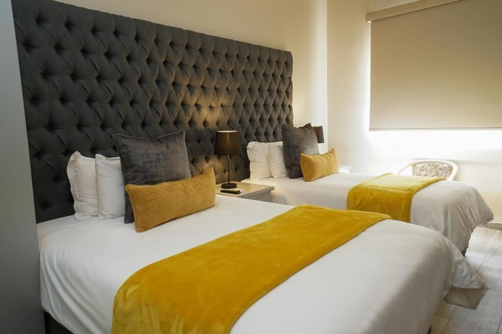 Habitación de hotel con dos camas, cabecera tapizada y mesita de noche con lámpara.