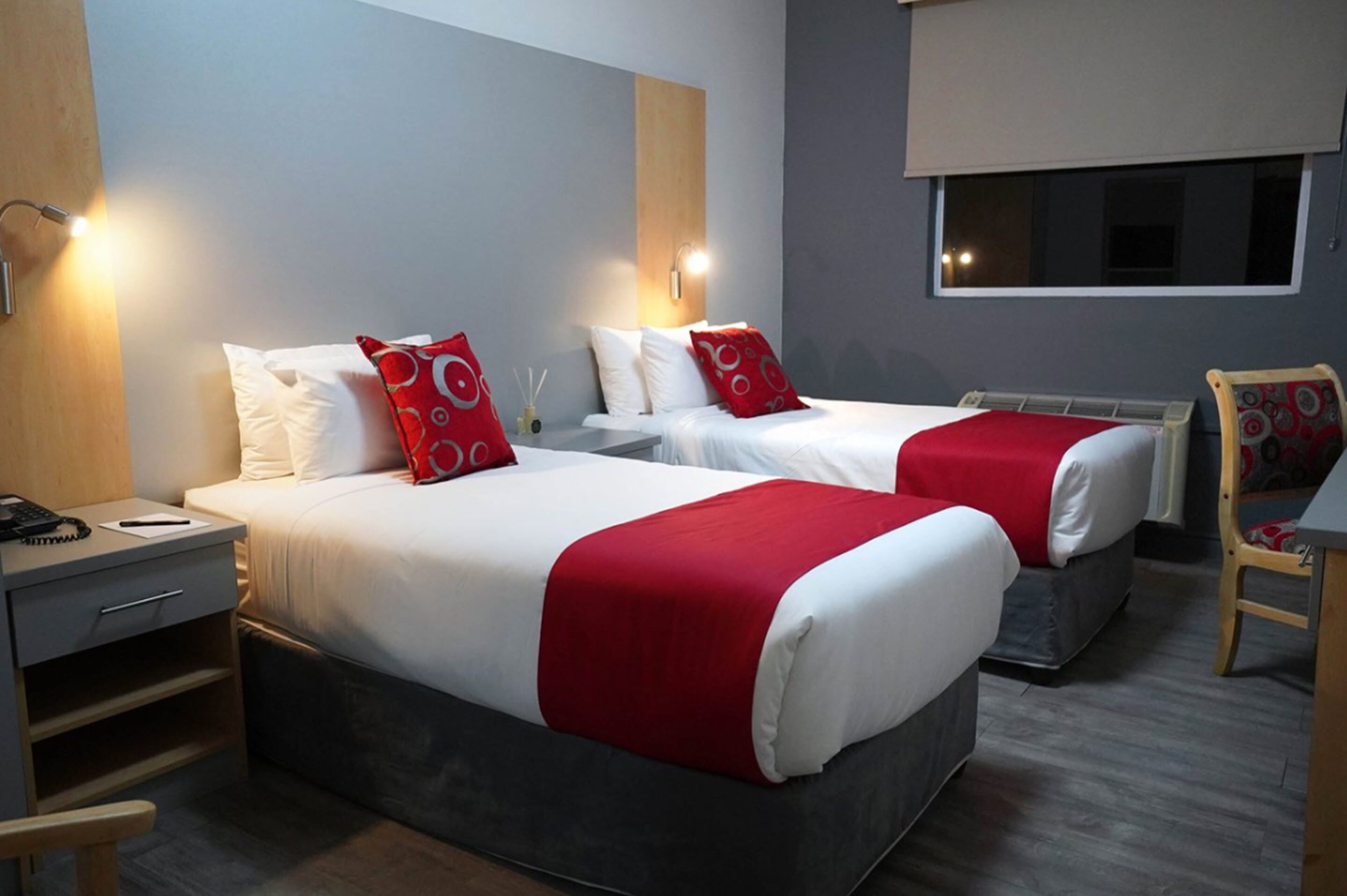 A hotel room with two beds, each with red and white bedding, a bedside table with a lamp, a chair, and a window with a view.
