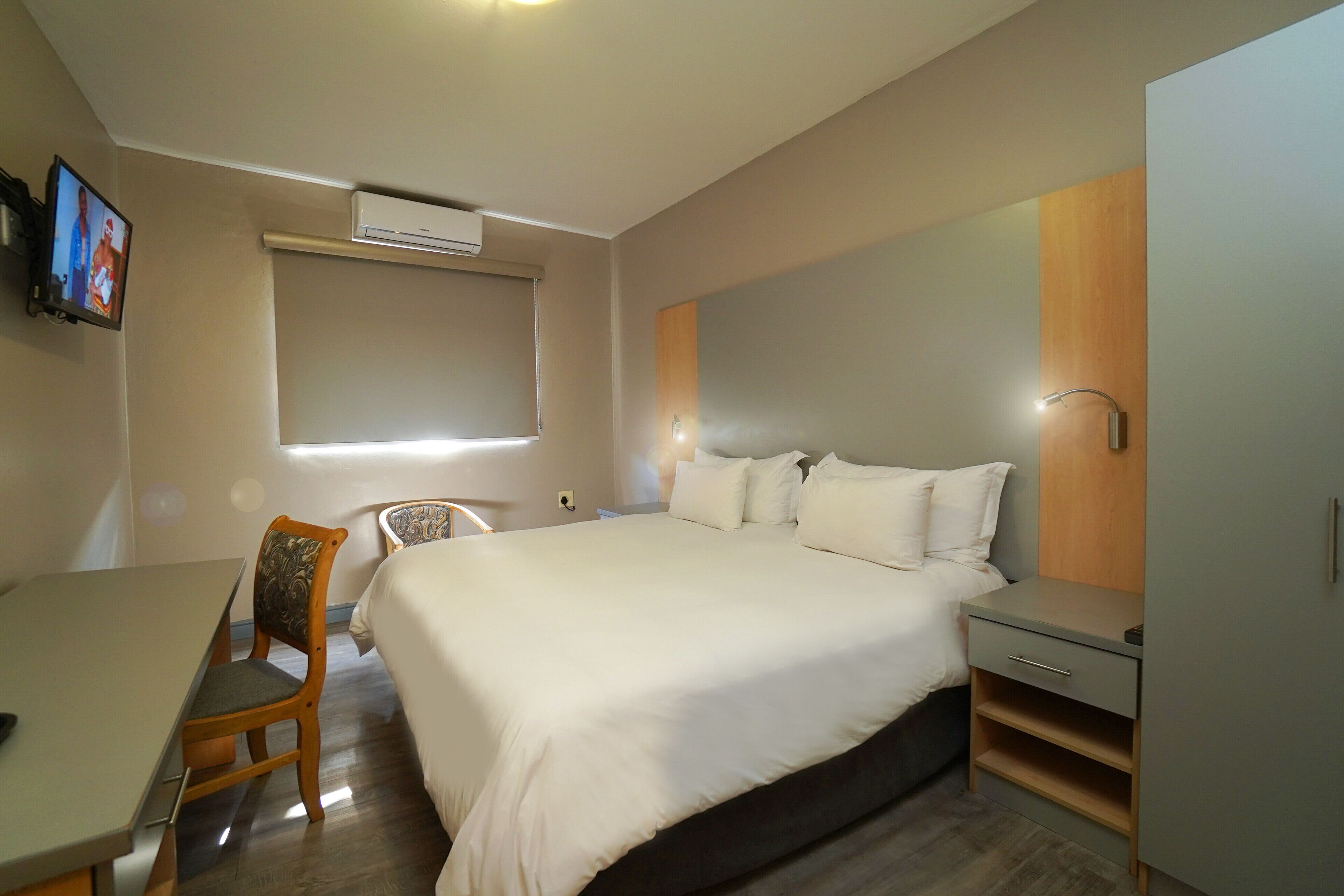 A hotel room with a bed, a desk, a chair, a TV, and an air conditioning unit.