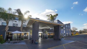 A hotel entrance with a covered walkway, palm trees, and a parking lot. - The Gallagher Hotel (Midrand)